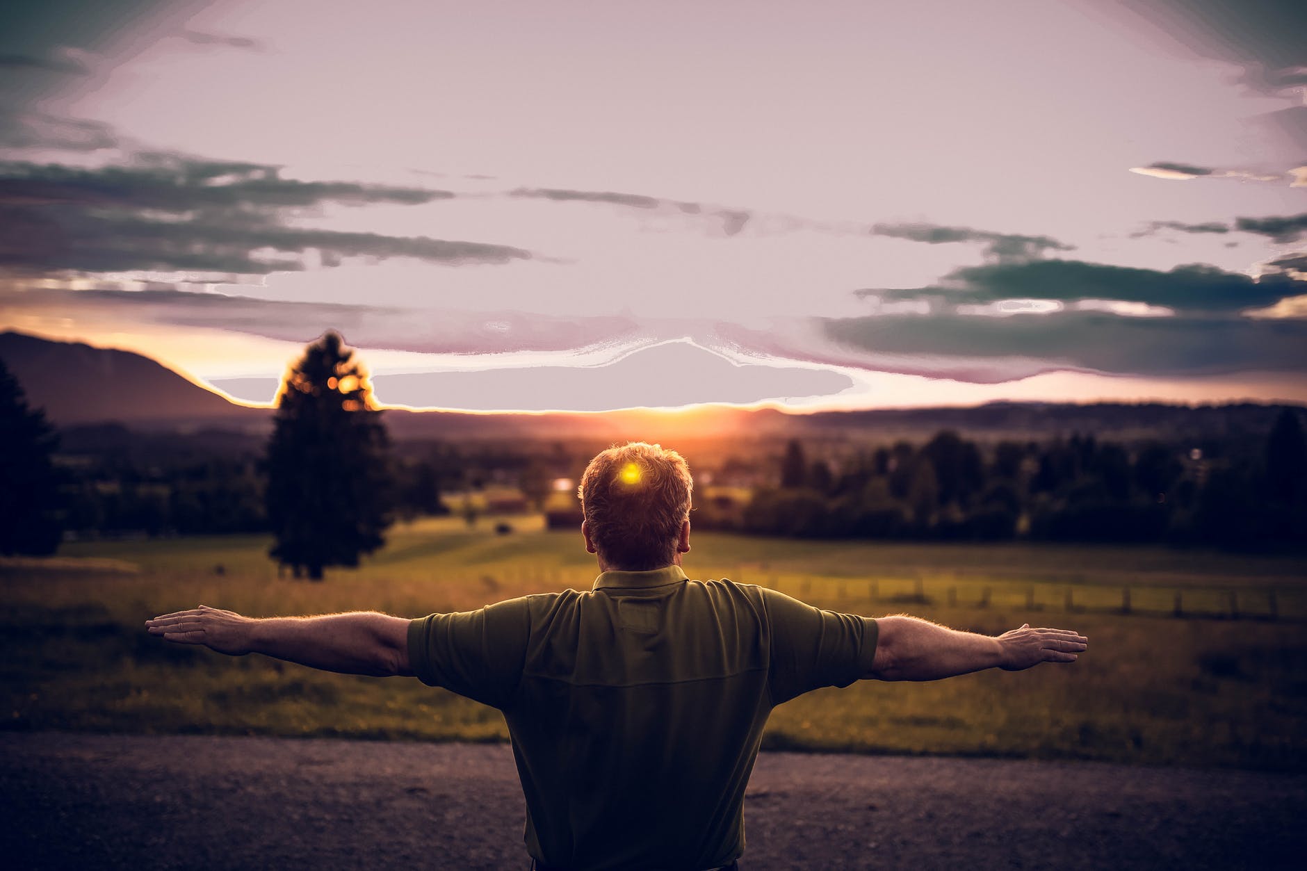 Man with outstretched arms looking at sun setting over fields