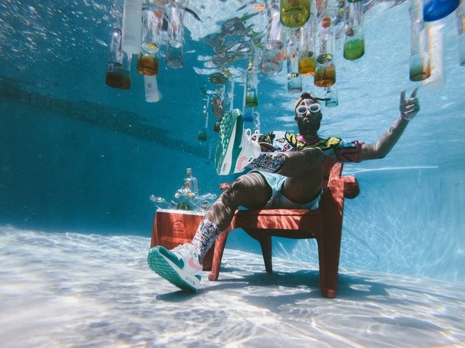 Man sitting on a chair at the bottom of a swimming pool surrounded by liquor bottles
