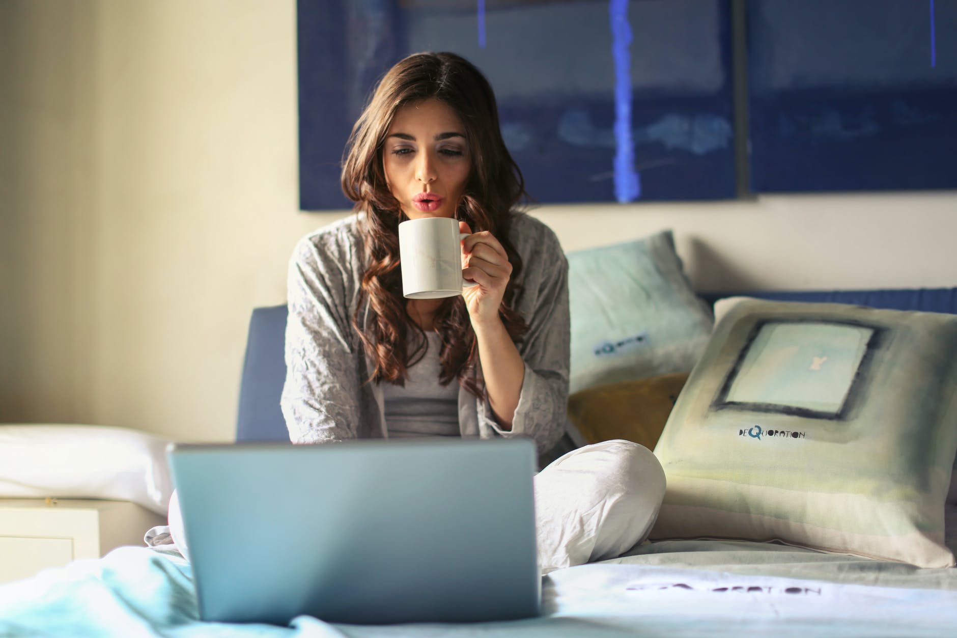 Woman blowing on a hot drink while sitting on her bed using a laptop