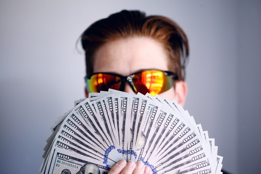 Man wearing mirrored sunglasses holding a fan of bank notes in front of his face