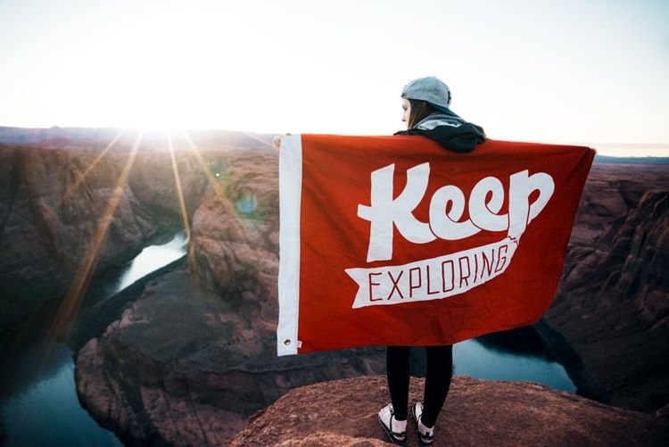 Woman on top of a cliff holding a banner that says 'keep exploring'