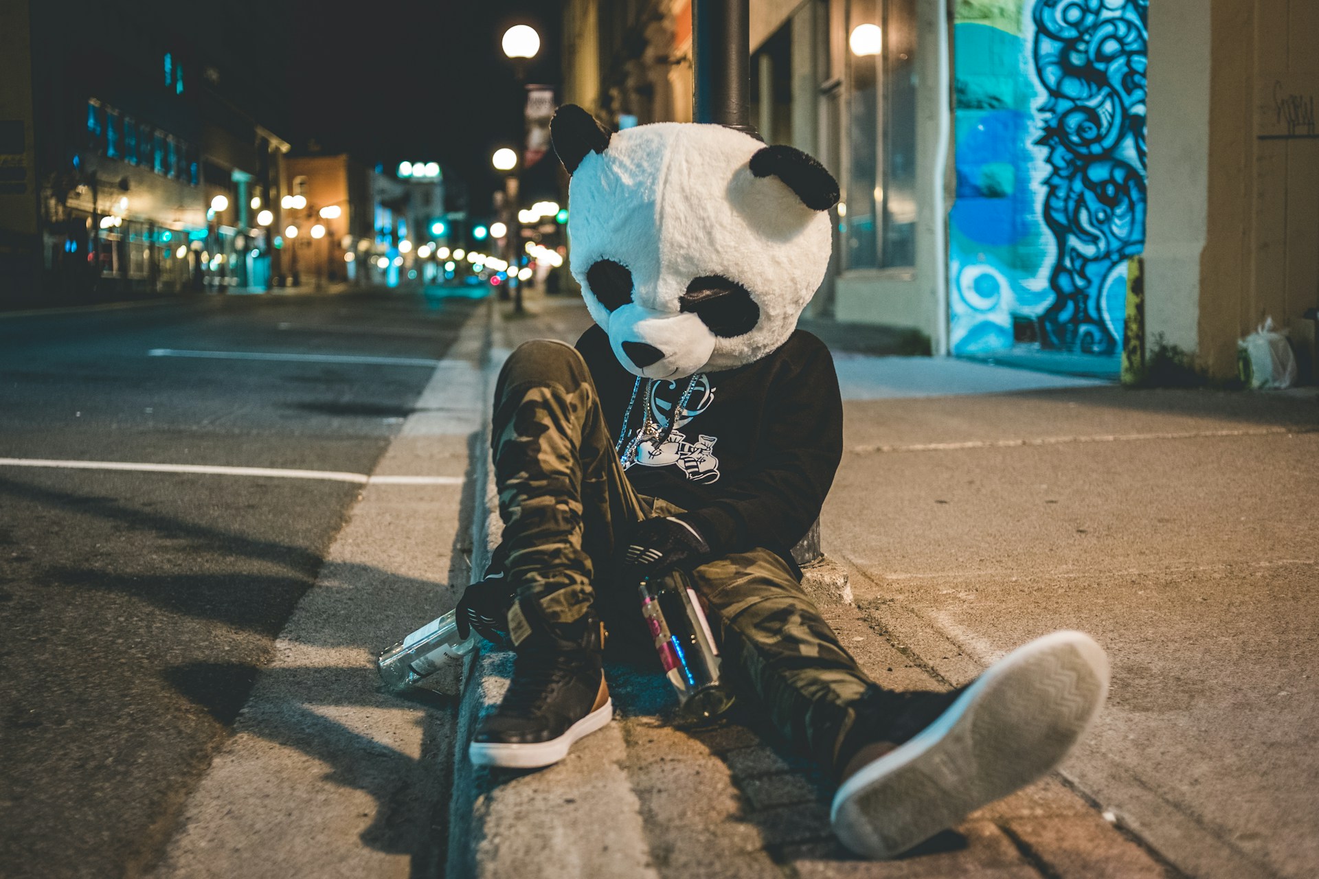 Person wearing a panda head sitting on the sidewalk clutching an empty bottle of alcohol