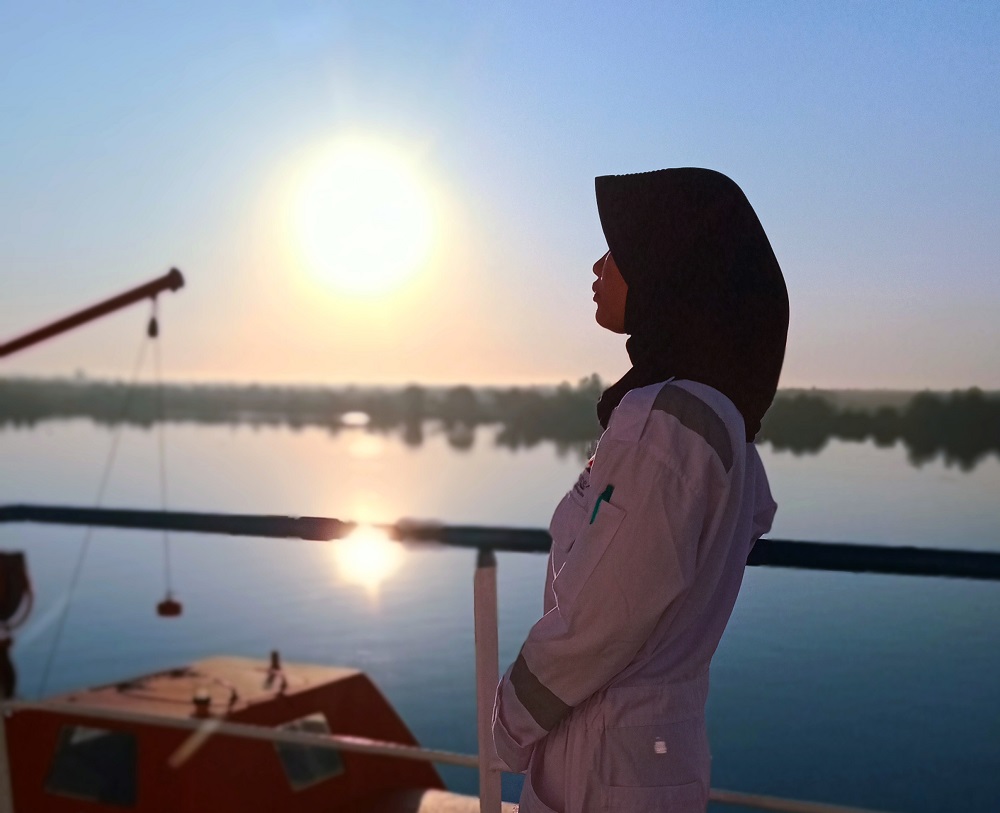 A hijab-wearing woman working in a seafarer job and standing on deck at sunset
