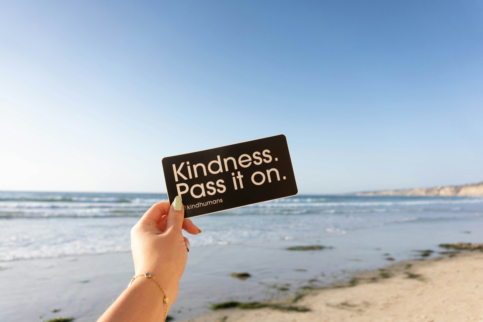 hand holding card saying 'Kindness: pass it on" in front of the ocean