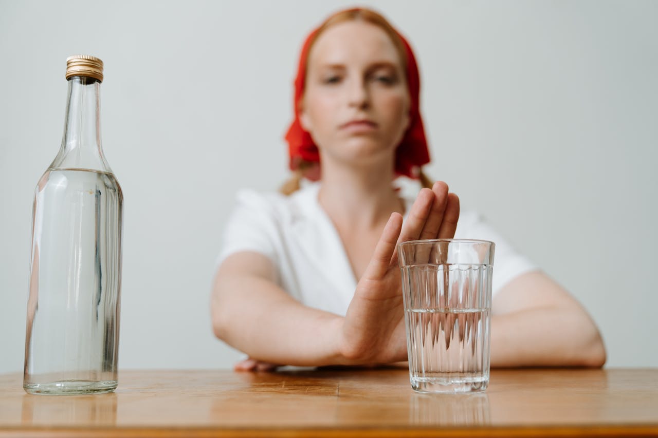 a woman sitting in front of a glass and bottle with her hand over the top of the glass