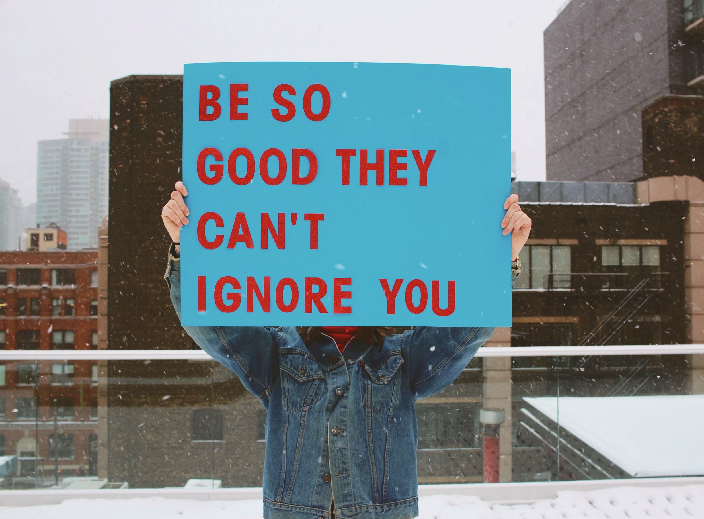 person holding a sign saying 'be so good they can't ignore you'